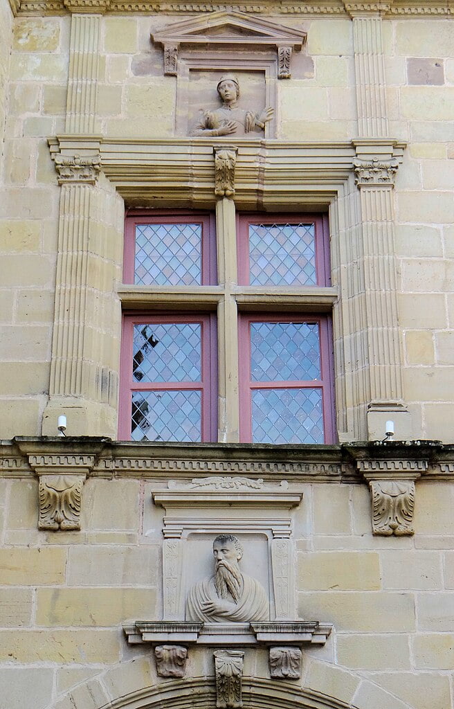 Stone building facade with two red-trimmed windows and two sculpted busts, one above and one below the windows.